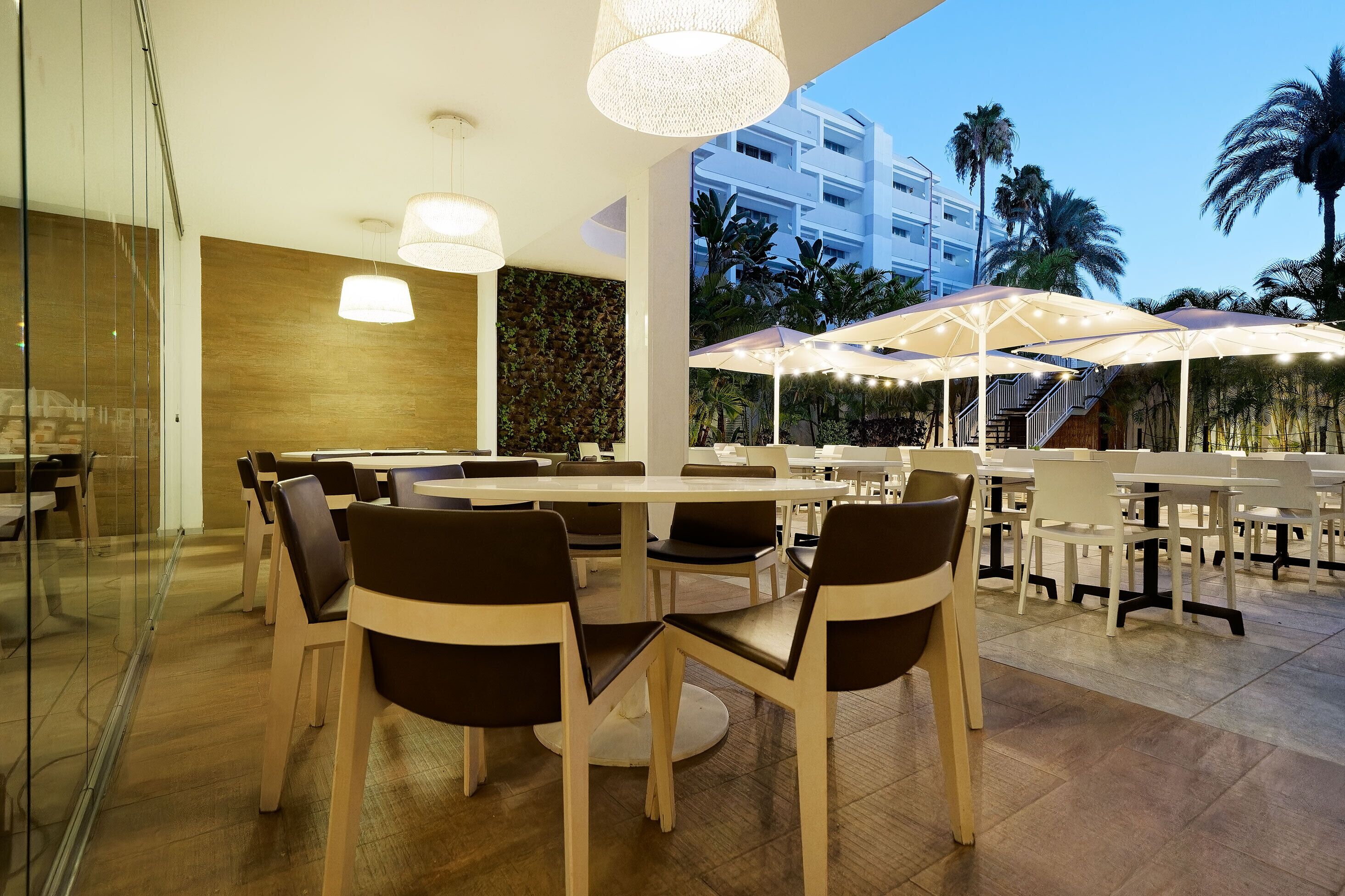 Een moderne eetruimte met ronde tafels en stoelen, parasols en uitzicht op palmbomen en een gebouw op de achtergrond.