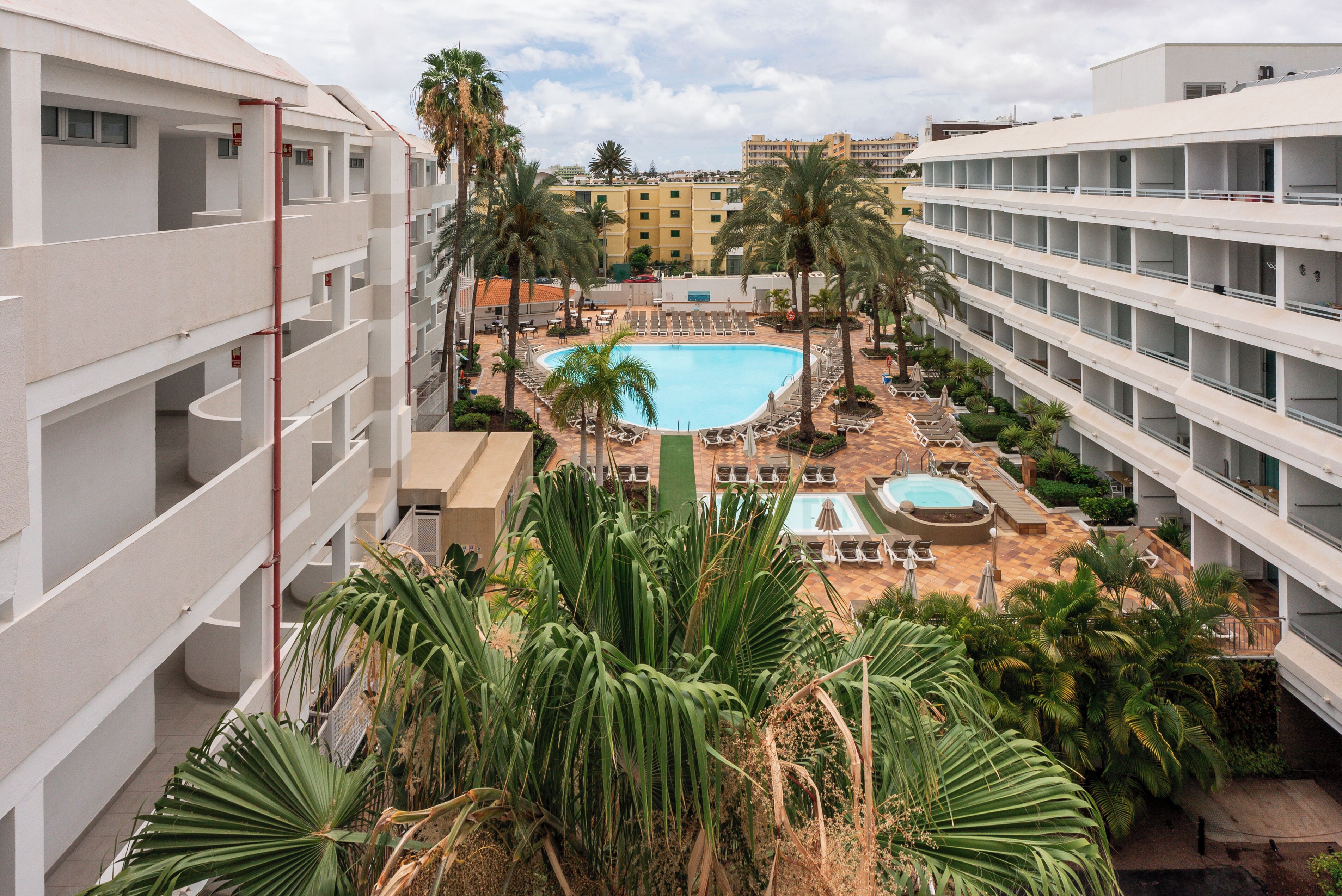Et hotel med flere etager, en central swimmingpool og palmer omkring.