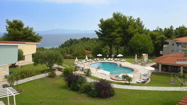 A pool area with lounge chairs and umbrellas, surrounded by greenery and a view of the sea. - Alkion Hotel (Kassandra)