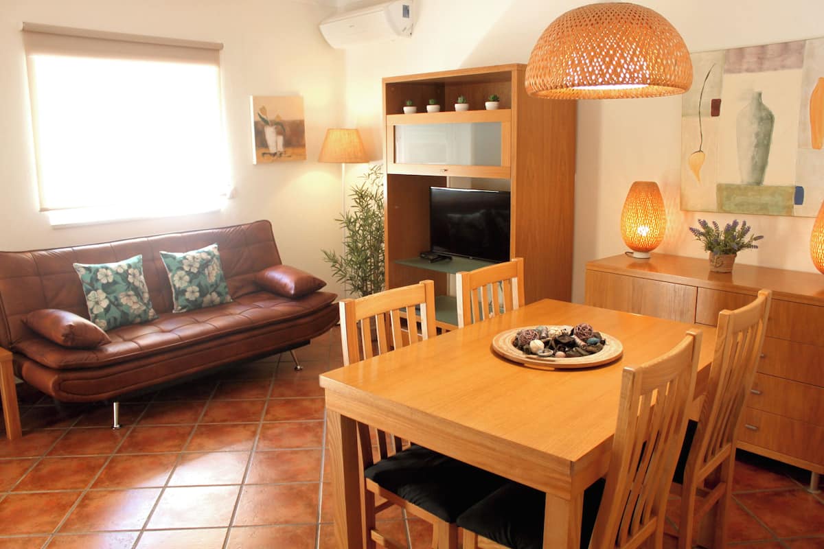 A living room with a brown leather sofa, a wooden dining table with chairs, a television, and a lamp.