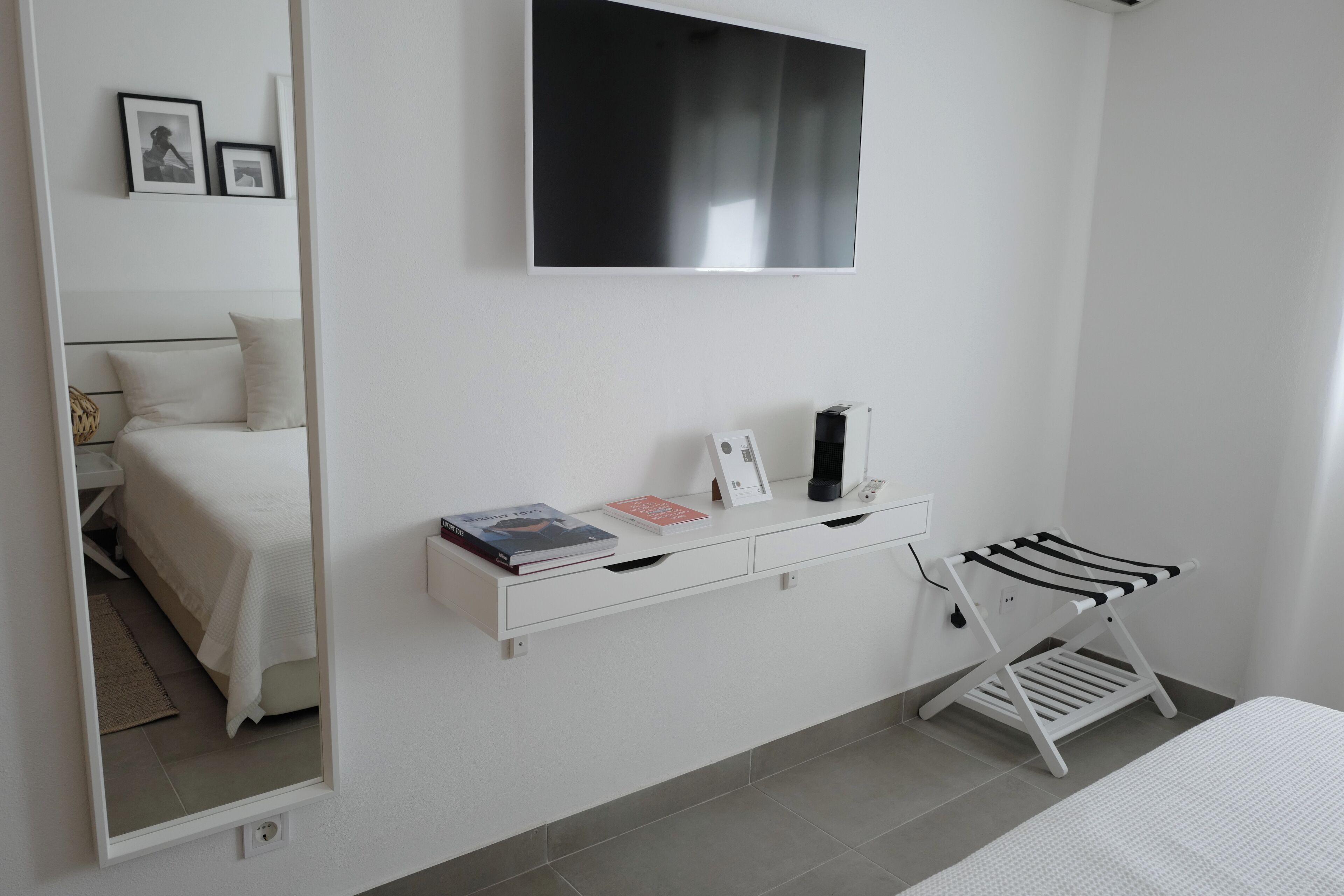 A modern bedroom with a bed, a bedside table with books and a lamp, a TV mounted on the wall, and a mirror reflecting the room.