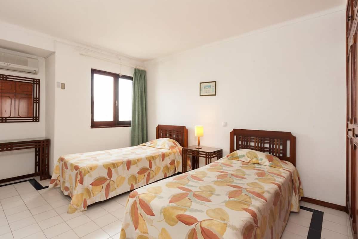 A hotel room with two single beds, a small table, a lamp, a window with curtains, and a wall-mounted air conditioner.