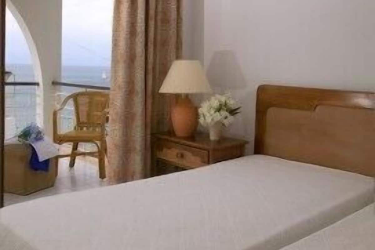 A hotel room with a bed, a nightstand with a lamp and flowers, a chair, and a view of the sea through an open door.