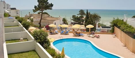 Une piscine dotée de chaises longues et de parasols, entourée de bâtiments et offrant une vue sur la mer.