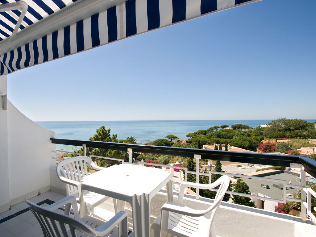 A balcony with a white table and chairs, overlooking a coastal view with trees and buildings.