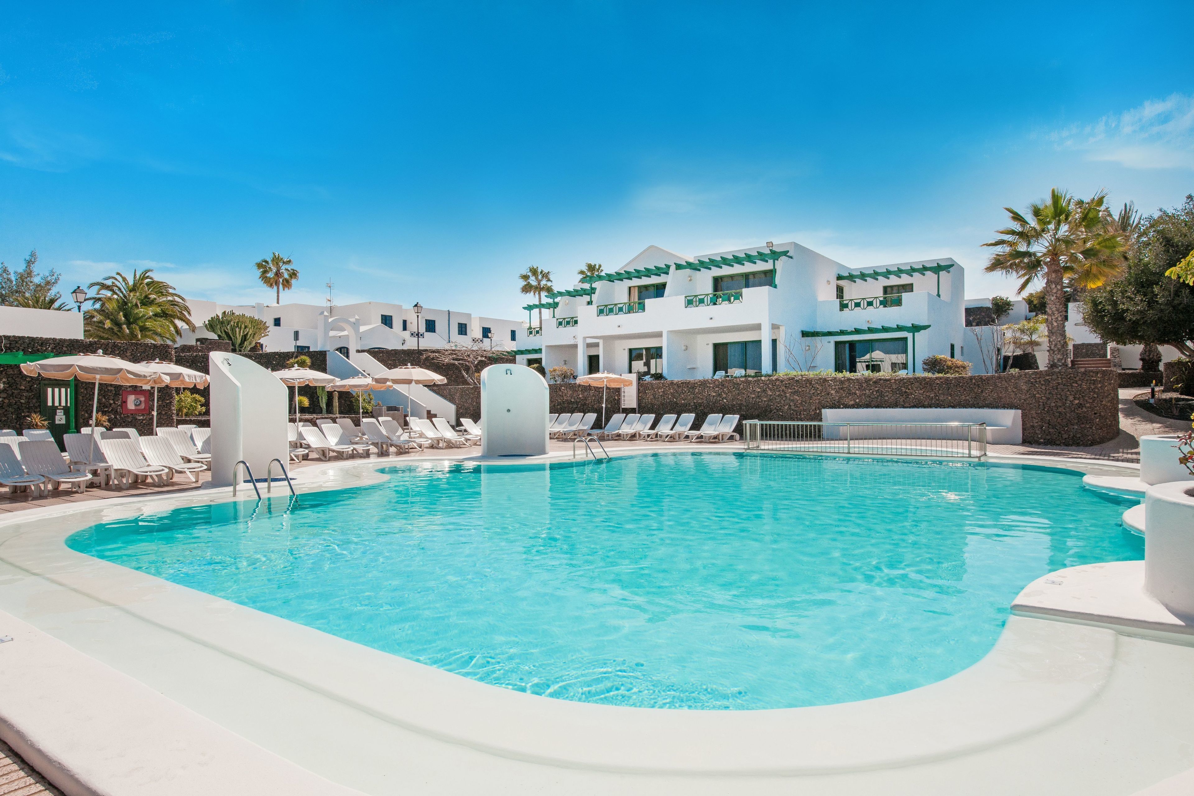 Una piscina con sillas de descanso y sombrillas, rodeada de edificios blancos con detalles verdes.