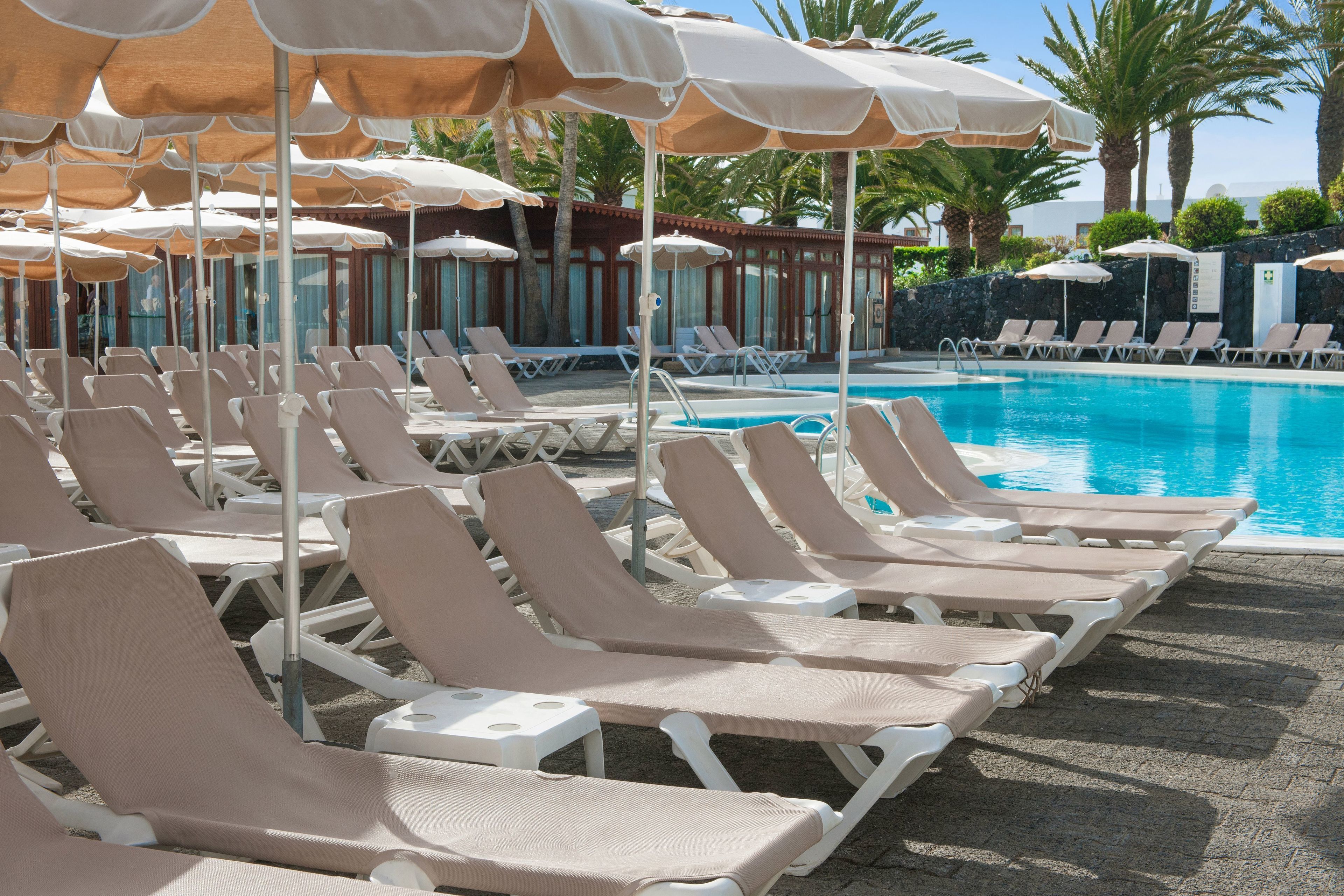 A poolside area with rows of sun loungers and umbrellas, a clear blue pool, and palm trees in the background.