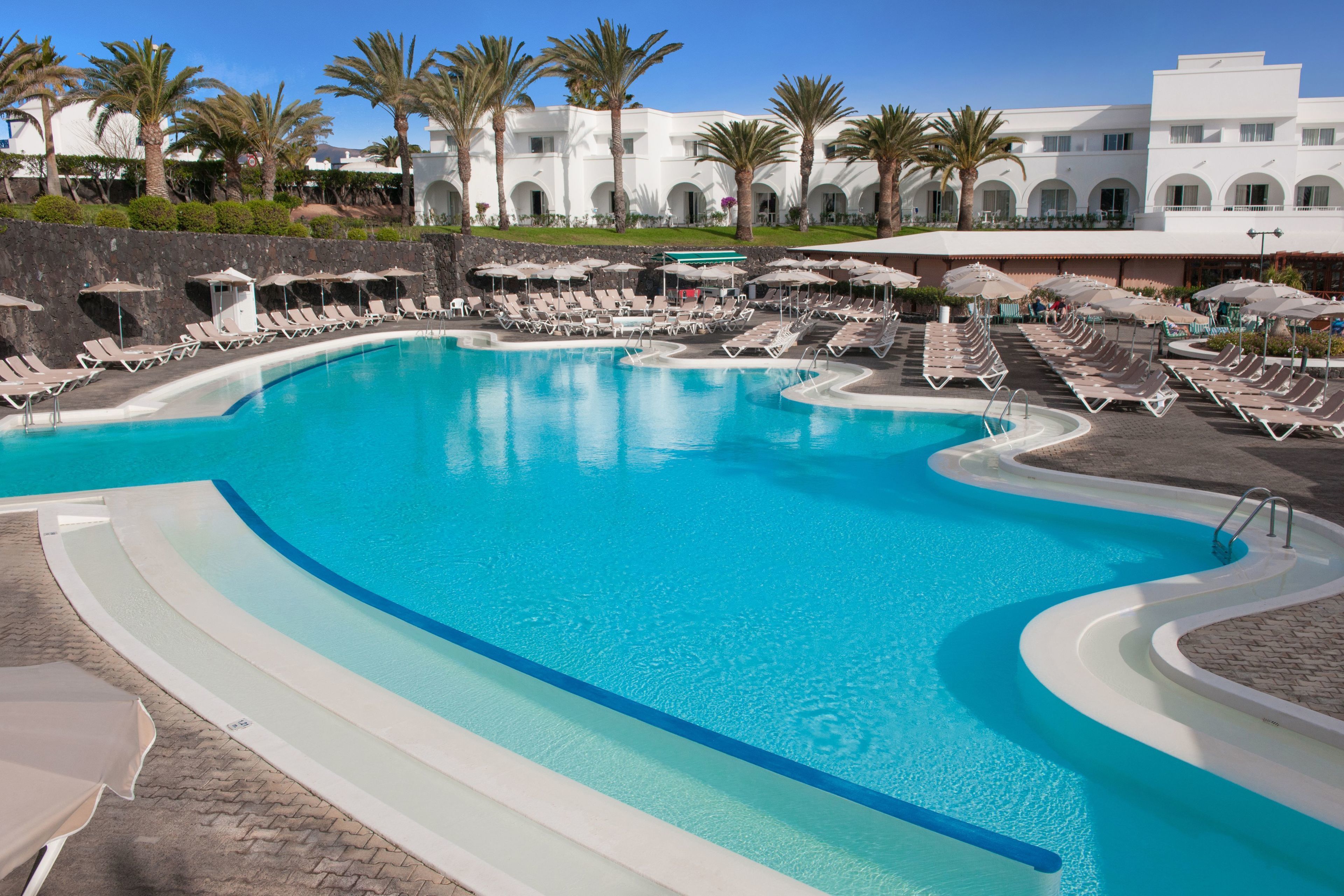 A large, curved swimming pool with multiple lounge areas and palm trees.