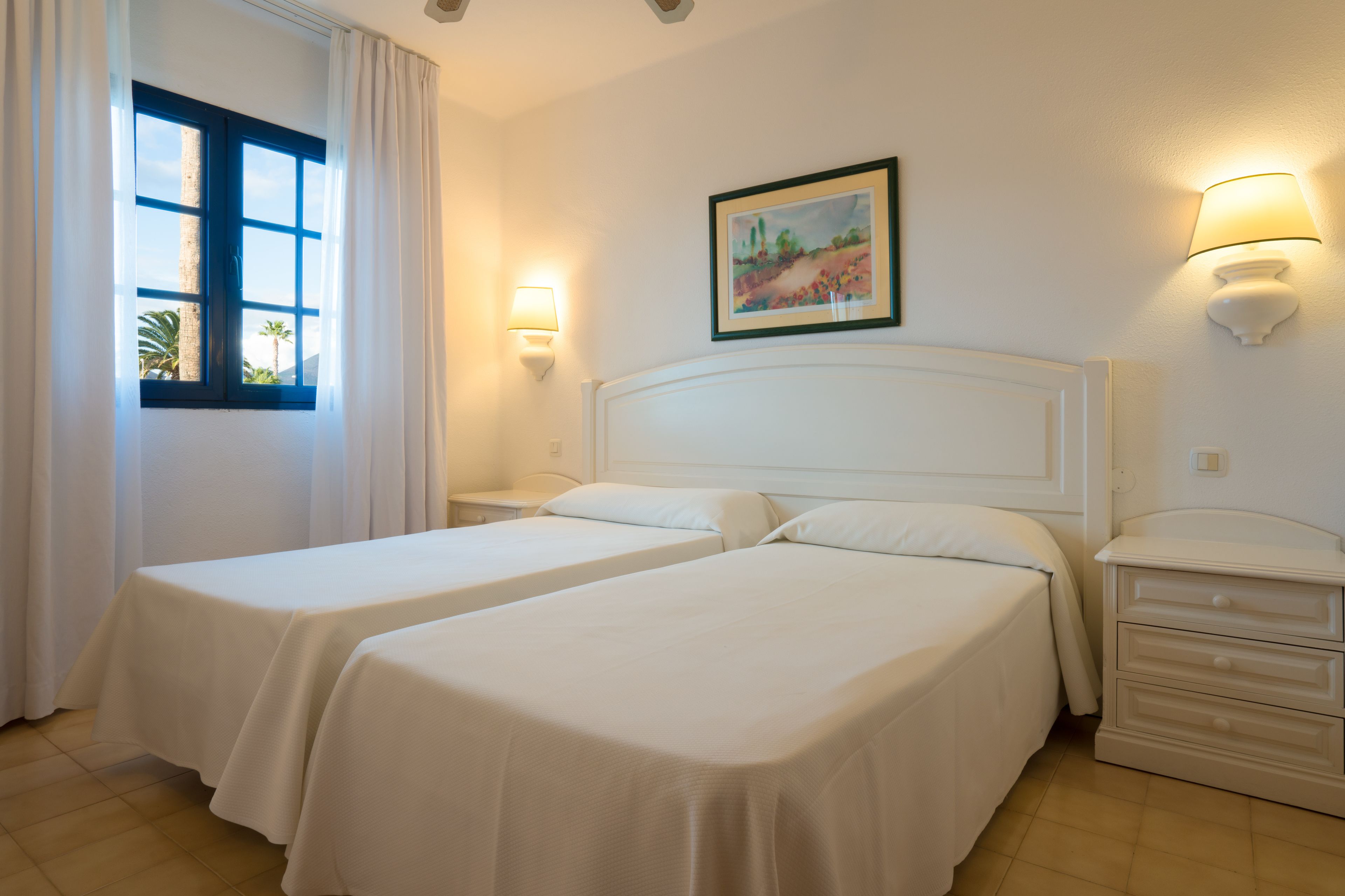 A hotel room with two beds, a nightstand, a lamp, and a painting on the wall.