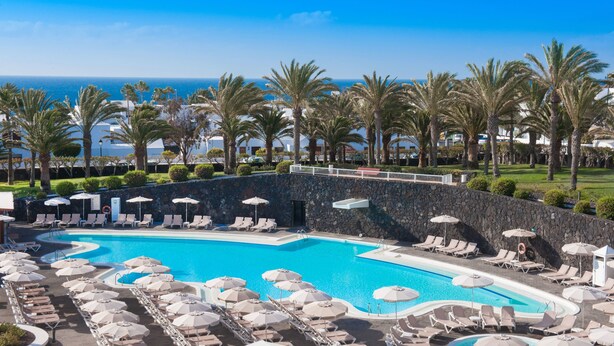 Zona de piscina en un resort con tumbonas y sombrillas, rodeada de palmeras y vistas al océano.