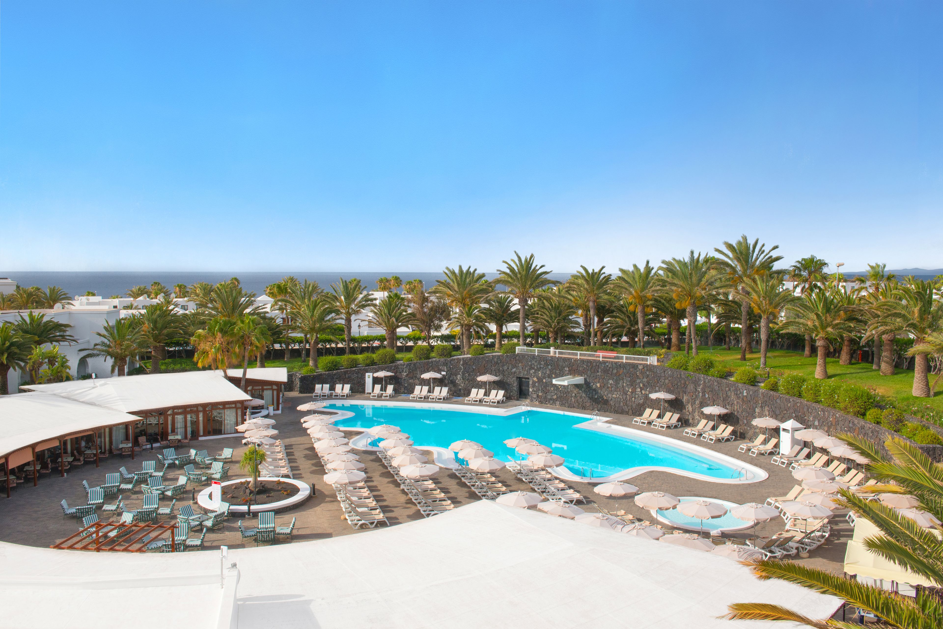 Zwembad van een resort met diverse ligbedden en parasols, omringd door palmbomen en uitzicht op de oceaan.