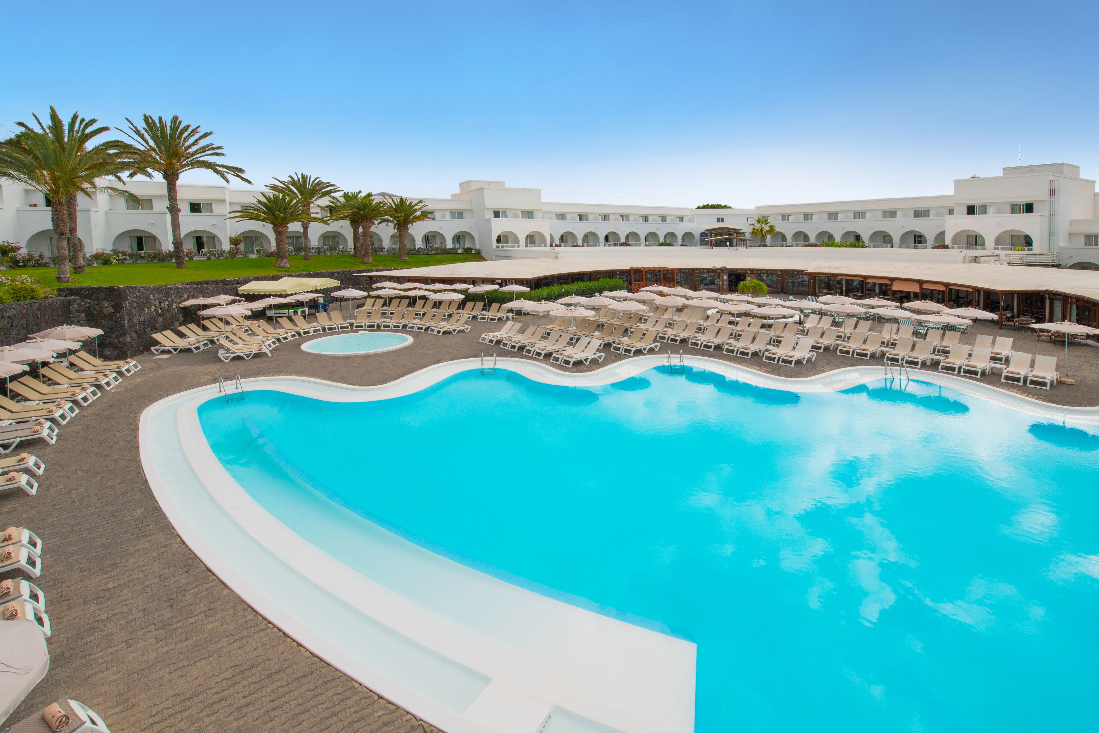 A large outdoor swimming pool surrounded by sun loungers and umbrellas, with a building in the background.