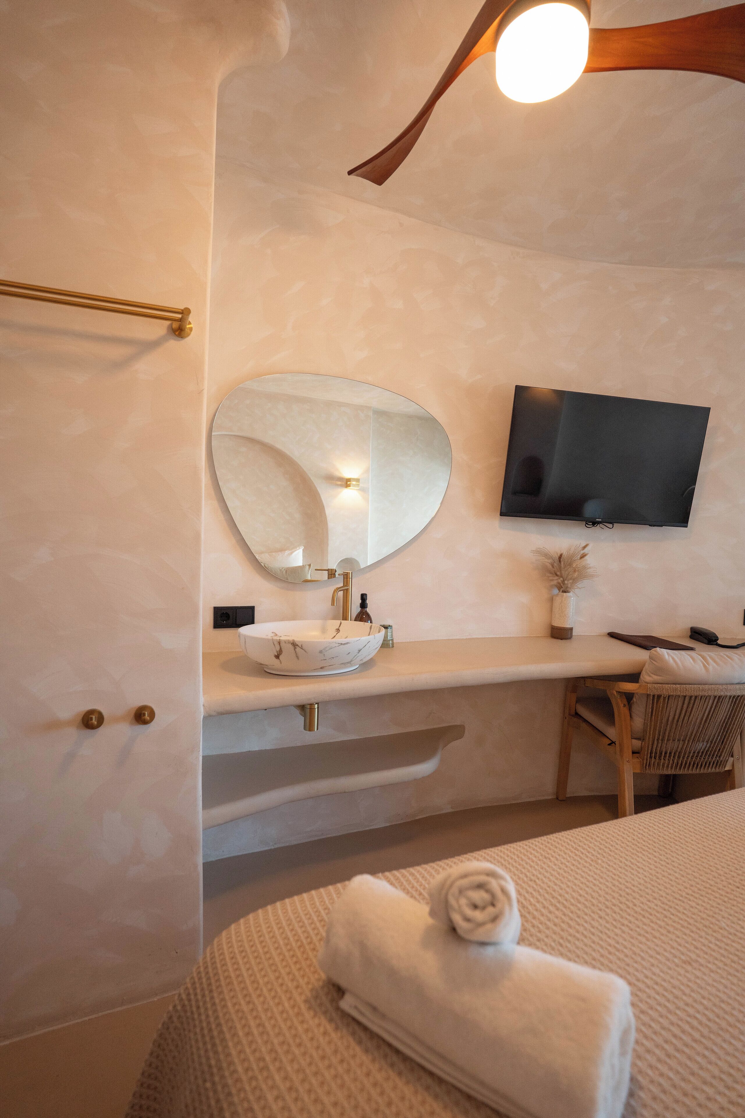 A modern bathroom with a round mirror, a sink, a flat-screen TV, and a towel on the bed.