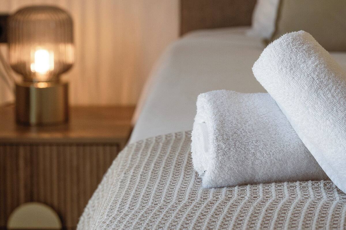 Una cama bien tendida con sábanas blancas y una toalla enrollada, una lámpara de noche y una mesita de madera con un espejo redondo.