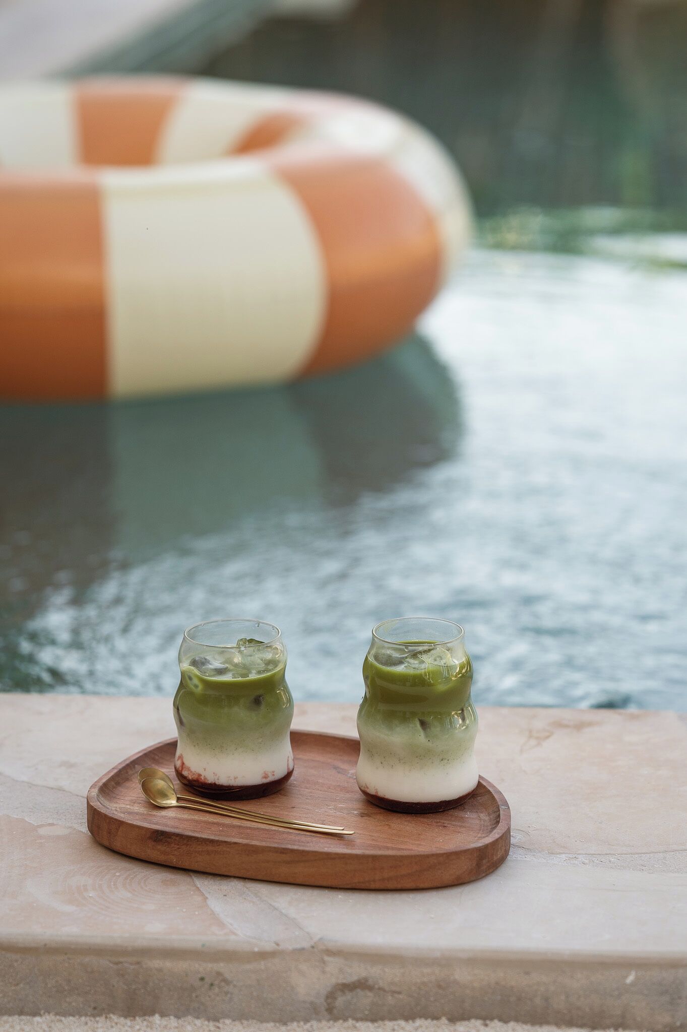 Two glasses of green beverage on a wooden tray by a pool with a lifebuoy in the background.