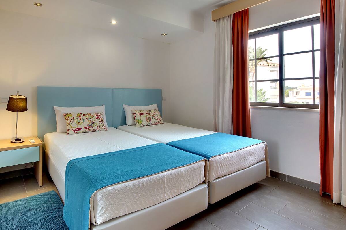 A double bed with two pillows, a bedside table with a lamp, and a window with curtains.