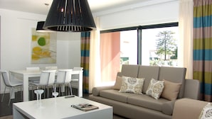 A modern living room with a white coffee table, a beige sofa with patterned cushions, and a dining area with white chairs and a white table. - Novochoro Apartments (Albufeira)
