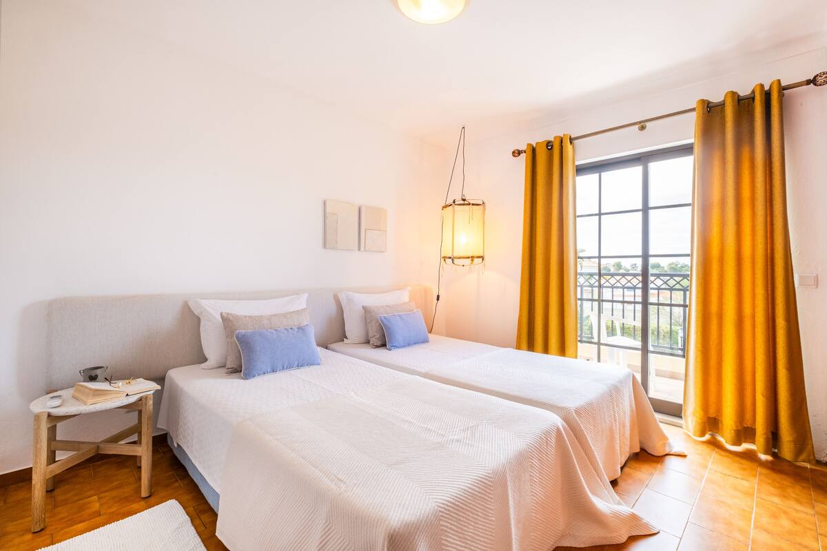 A hotel room with two beds, a nightstand, a lamp, and a window with curtains.