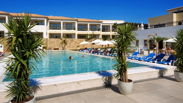 A pool area with lounge chairs and palm trees, surrounded by a building with multiple balconies. - Novochoro Apartments (Albufeira)