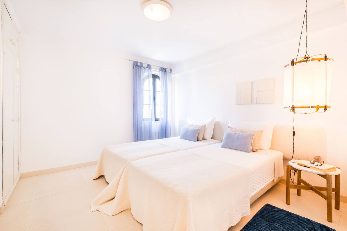 A bedroom with two beds, a window with curtains, a bedside table with a lamp, and a wall-mounted light fixture.