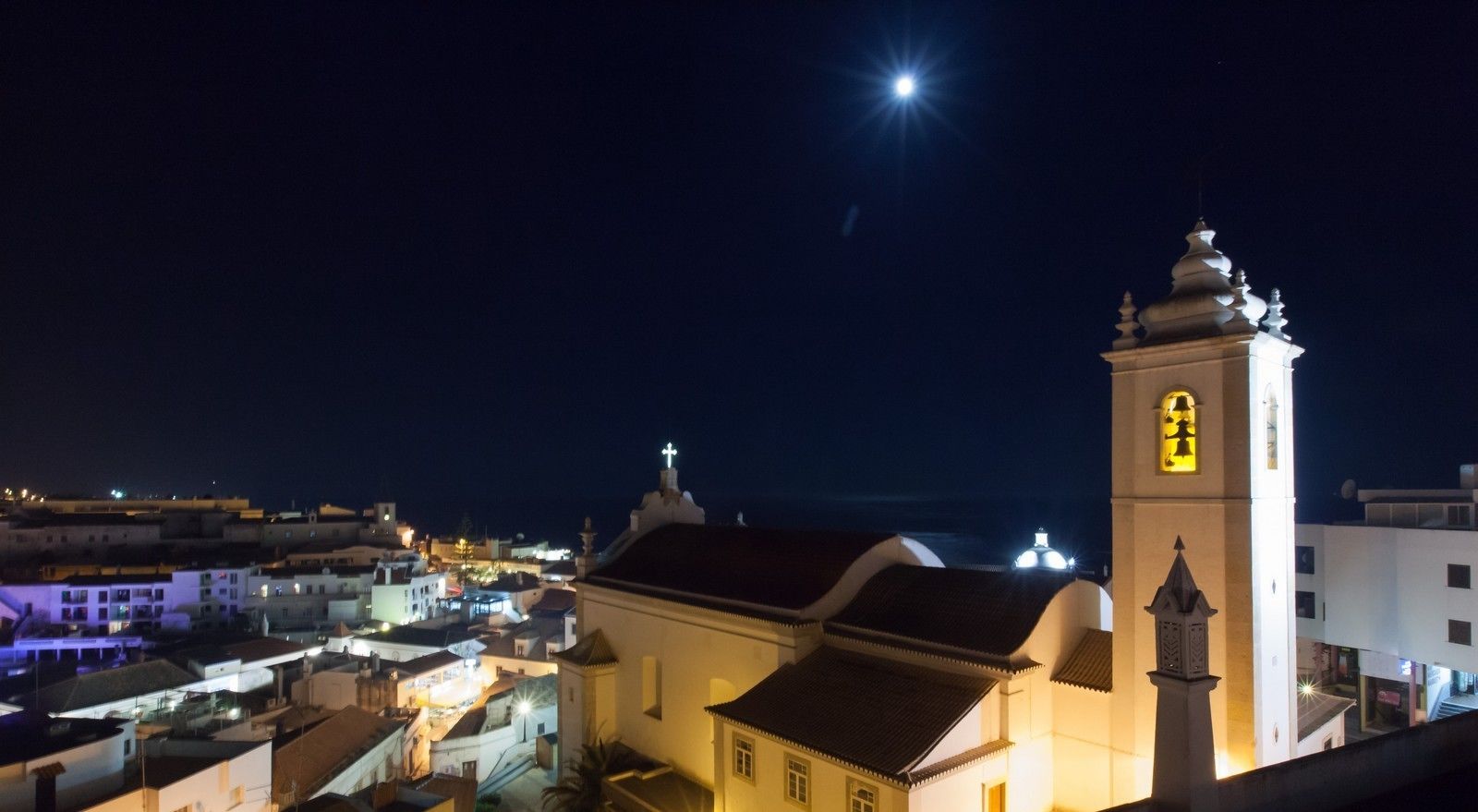 Ein Nachtblick auf eine Küstenstadt mit einer markanten Kirche, einem Glockenturm und einem Kreuz auf der Spitze.