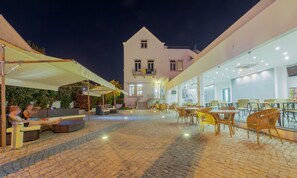 Uma área externa de café com assentos e mesas, um edifício com varanda ao fundo e um calçadão.