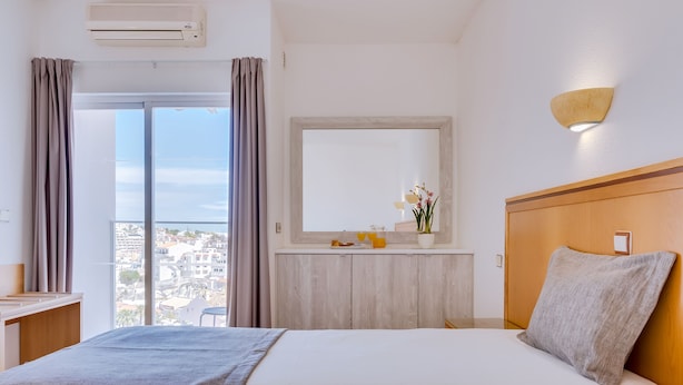 A hotel room with a large bed, a desk, a window with curtains, a mirror, and a wall-mounted light fixture. - Hotel Vila Recife (Albufeira)