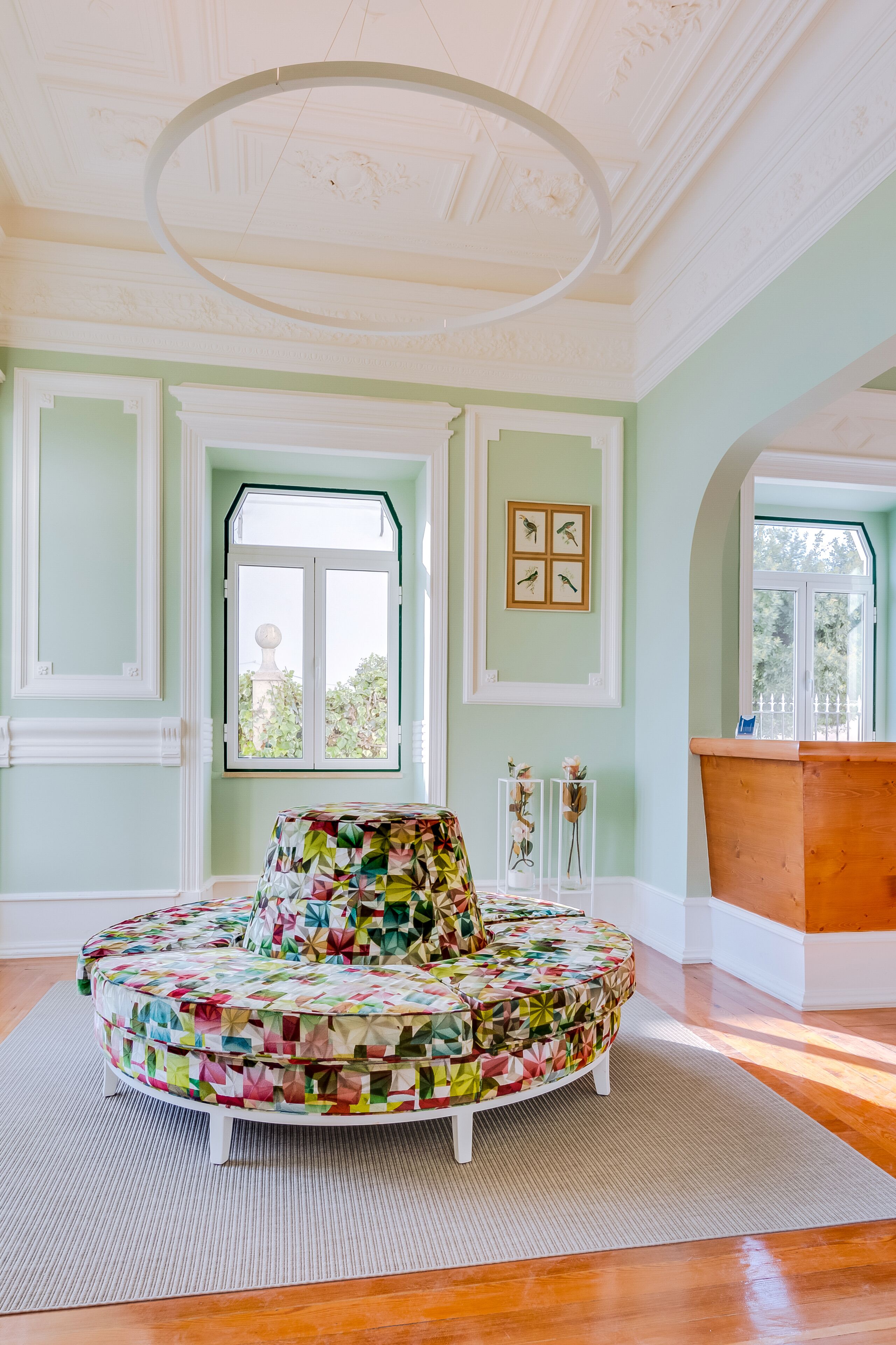 Ein Zimmer mit einem runden, bunten Ottomanen, einer Holzbank, einem großen Fenster und einer Decke mit dekorativer Stuckleiste.