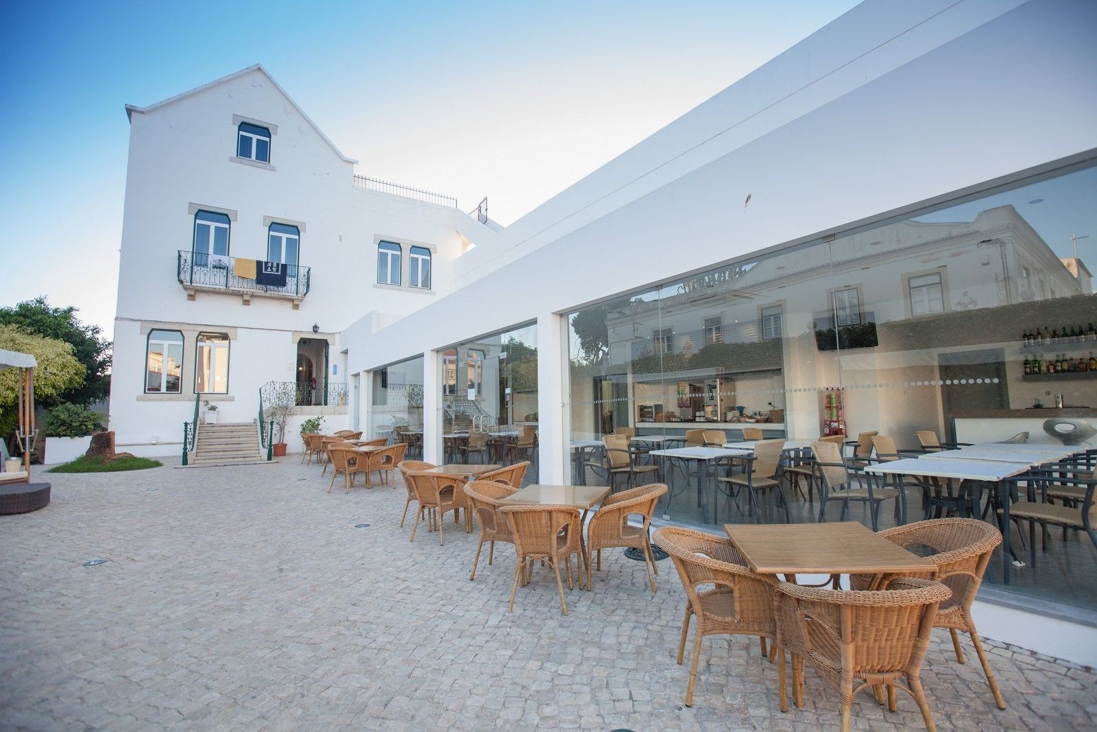 A building with a white exterior, outdoor seating area with wicker chairs and tables, and a glass facade reflecting the interior.