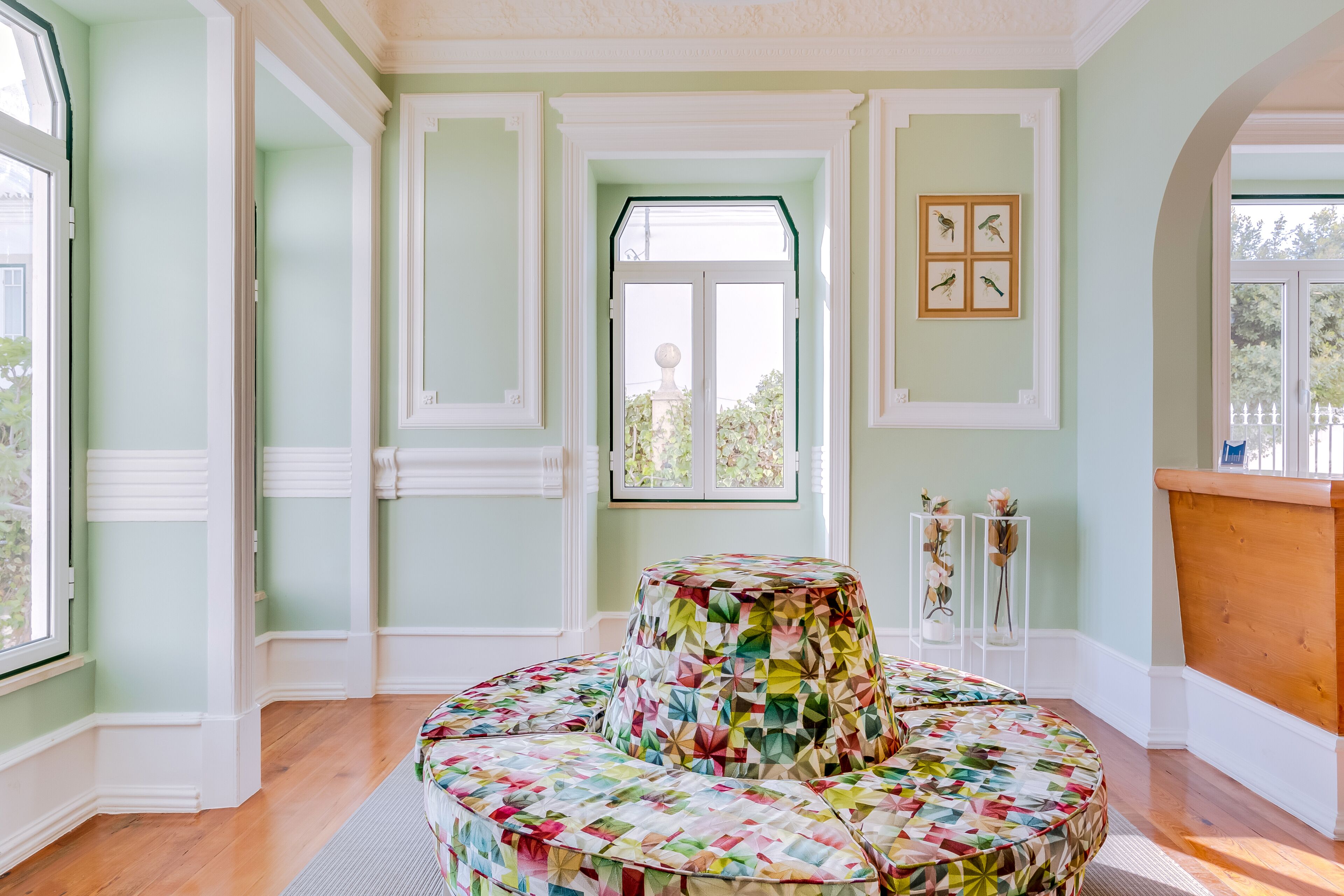 Una habitación con un otomanillo redondo, de varios niveles y colorido, acolchado en el centro, un ventanal con vistas a la vegetación y un cuadro con pájaros en la pared.