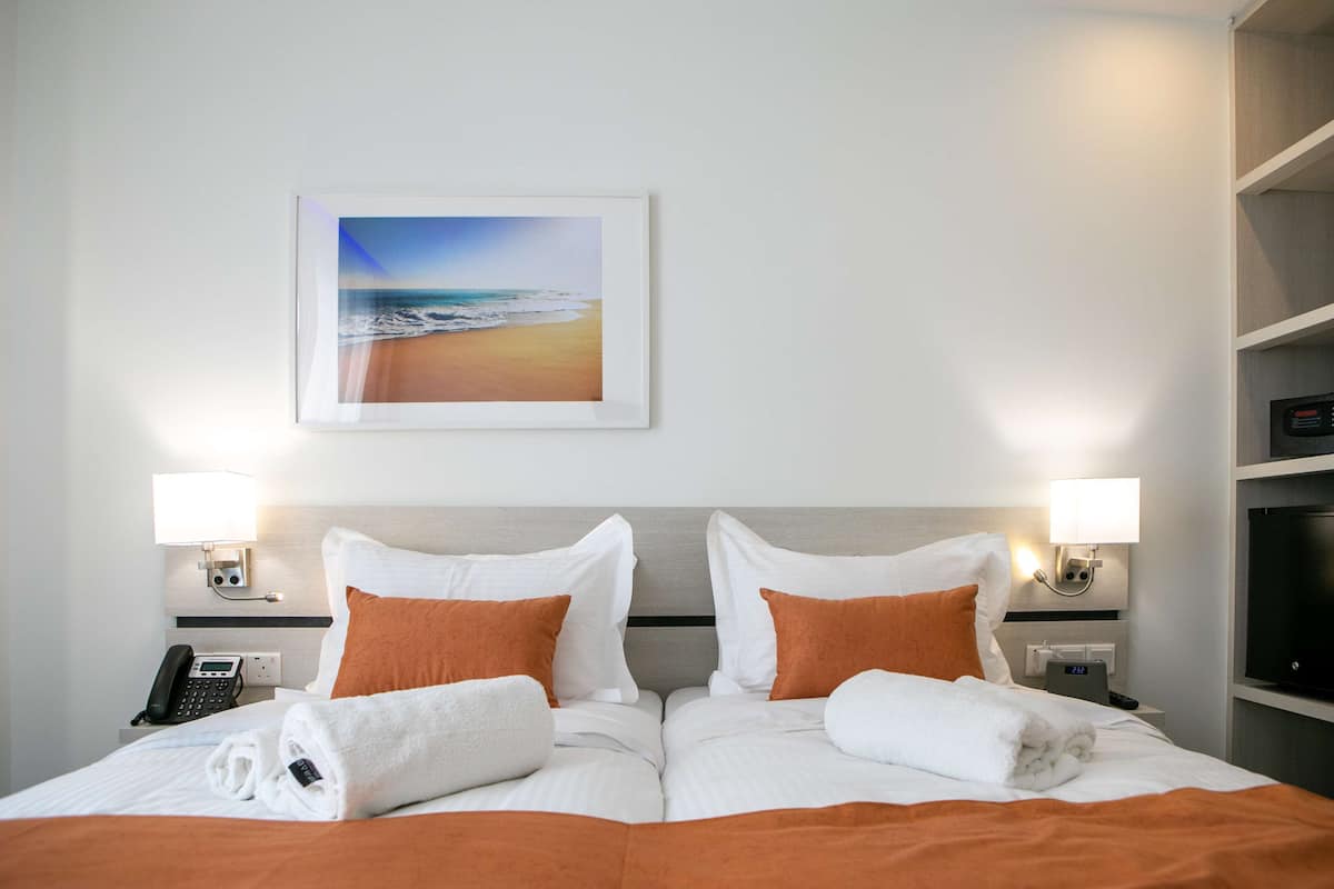Ein Hotelzimmer mit einem Bett, zwei Nachttischlampen, einem Strandbild, einem Telefon und einem Fernseher auf einem Regal.