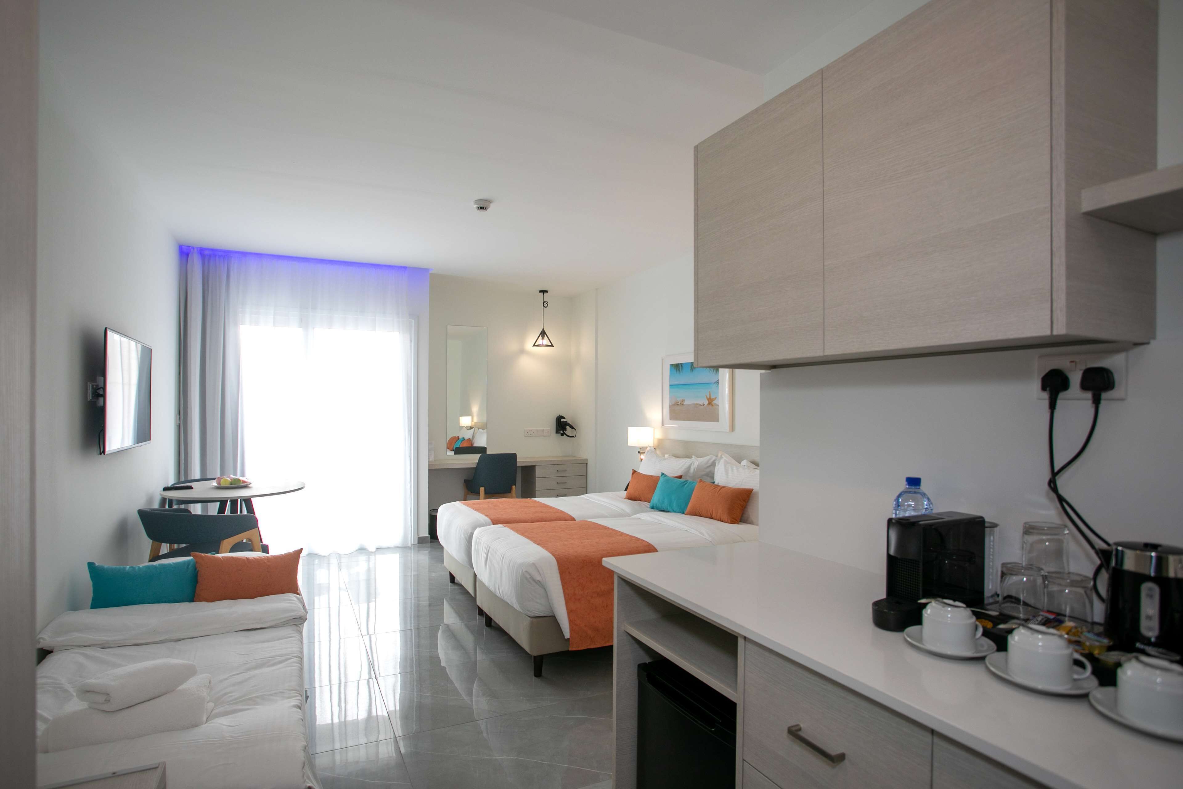A hotel room with a bed, a desk, a chair, a small table, a coffee maker, and a water bottle.