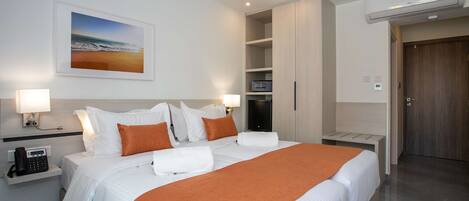 A hotel room with a bed, two bedside lamps, a painting of a beach, a wardrobe, a desk with a phone, and a door.