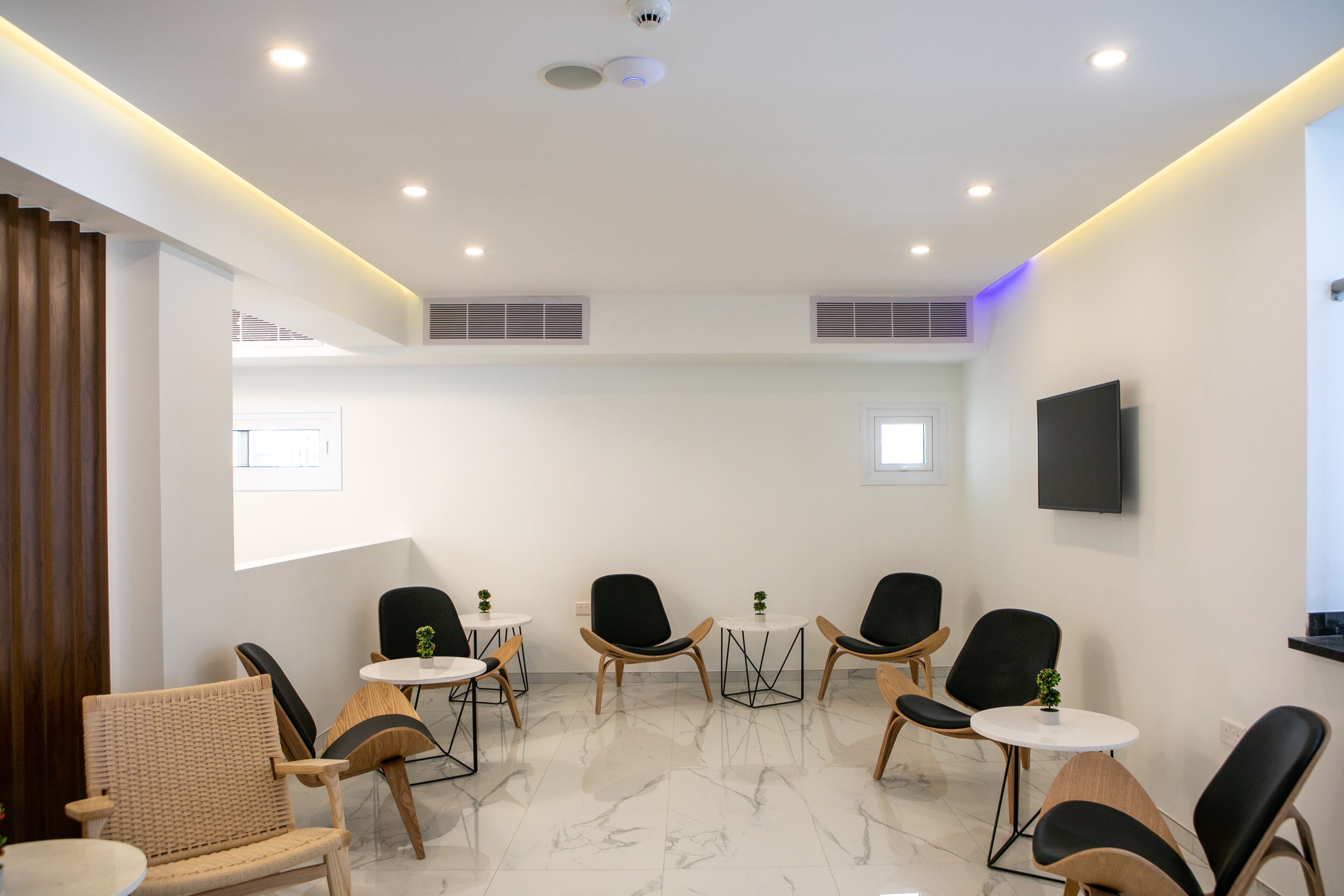 A modern waiting area with a mix of wicker and black chairs, small tables, and a flat-screen TV mounted on the wall.