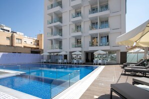 A rooftop swimming pool with lounge chairs and umbrellas, surrounded by a modern building with balconies.