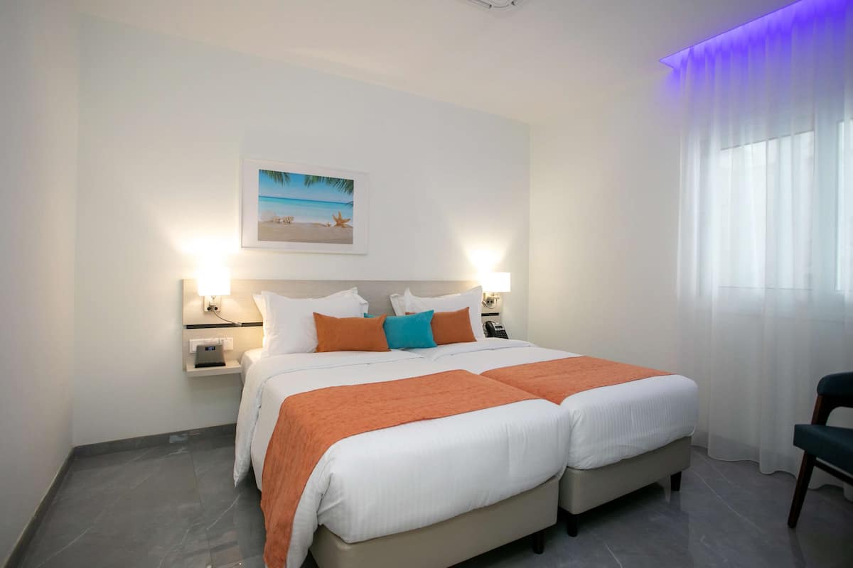 Ein Hotelzimmer mit einem großen Bett, zwei Nachttischen, einem Schminktisch und einem Bild in einem Rahmen an der Wand, sowie einem Fenster mit transparenten Vorhängen.