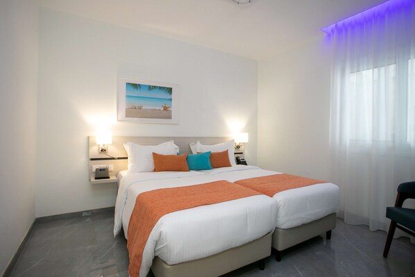 A hotel room with a large bed, two bedside tables, a nightstand, a framed picture on the wall, and a window with sheer curtains.