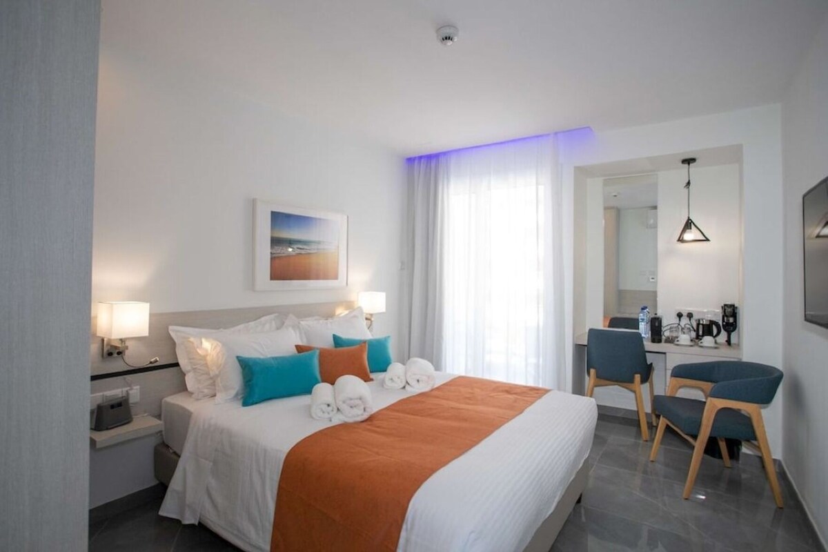 Habitación de hotel con cama, mesitas de noche, escritorio, sillas, espejo y un cuadro en la pared.