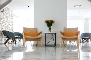 A modern lobby with two wicker chairs, a small table with a vase of flowers, and a white wall with a geometric pattern.
