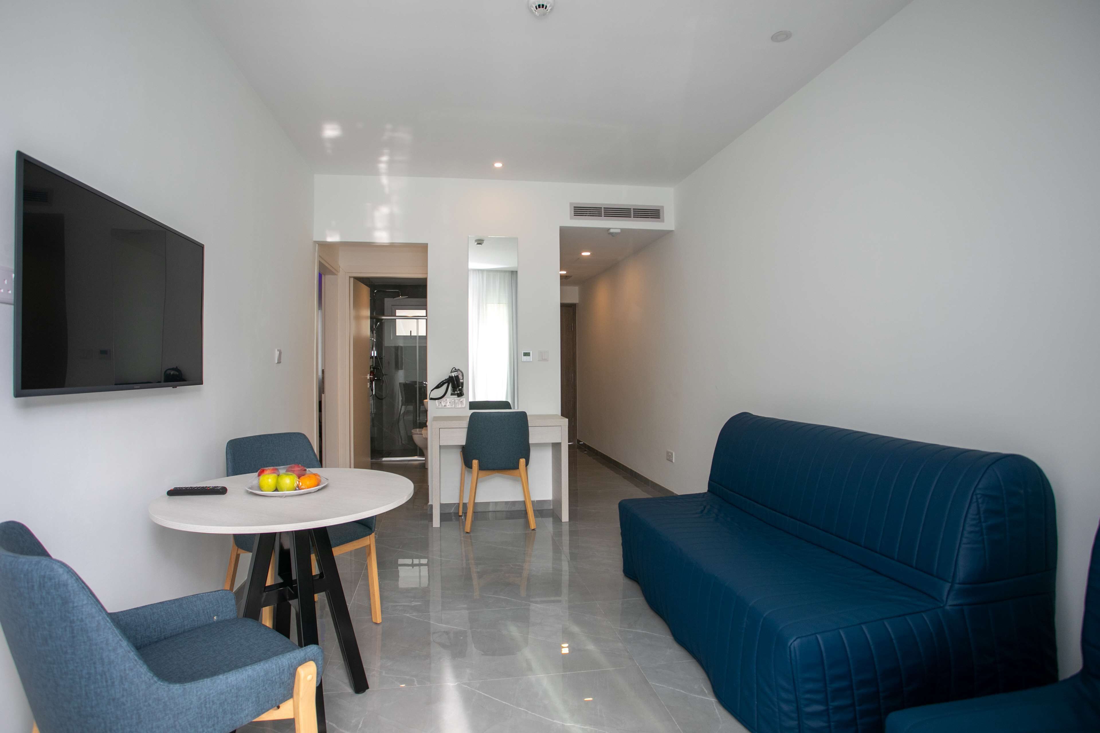 A modern living room with a blue sofa, a dining table with chairs, a flat-screen TV mounted on the wall, and a small kitchen area in the background.