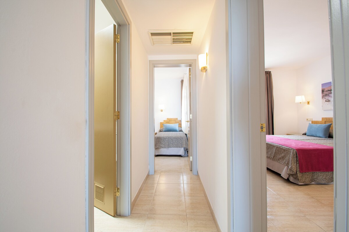 Un couloir menant à deux chambres à coucher, chacune équipée d’un lit, d’une bouche d’aération au plafond et d’un luminaire fixé au mur.