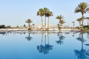 Une piscine bordée de palmiers et agrémentée de parasols, dont le reflet se mire dans l’eau.