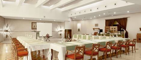 Een ruime balzaal met lange tafels gedekt voor een diner, een podium en een groot raam met gordijnen.
