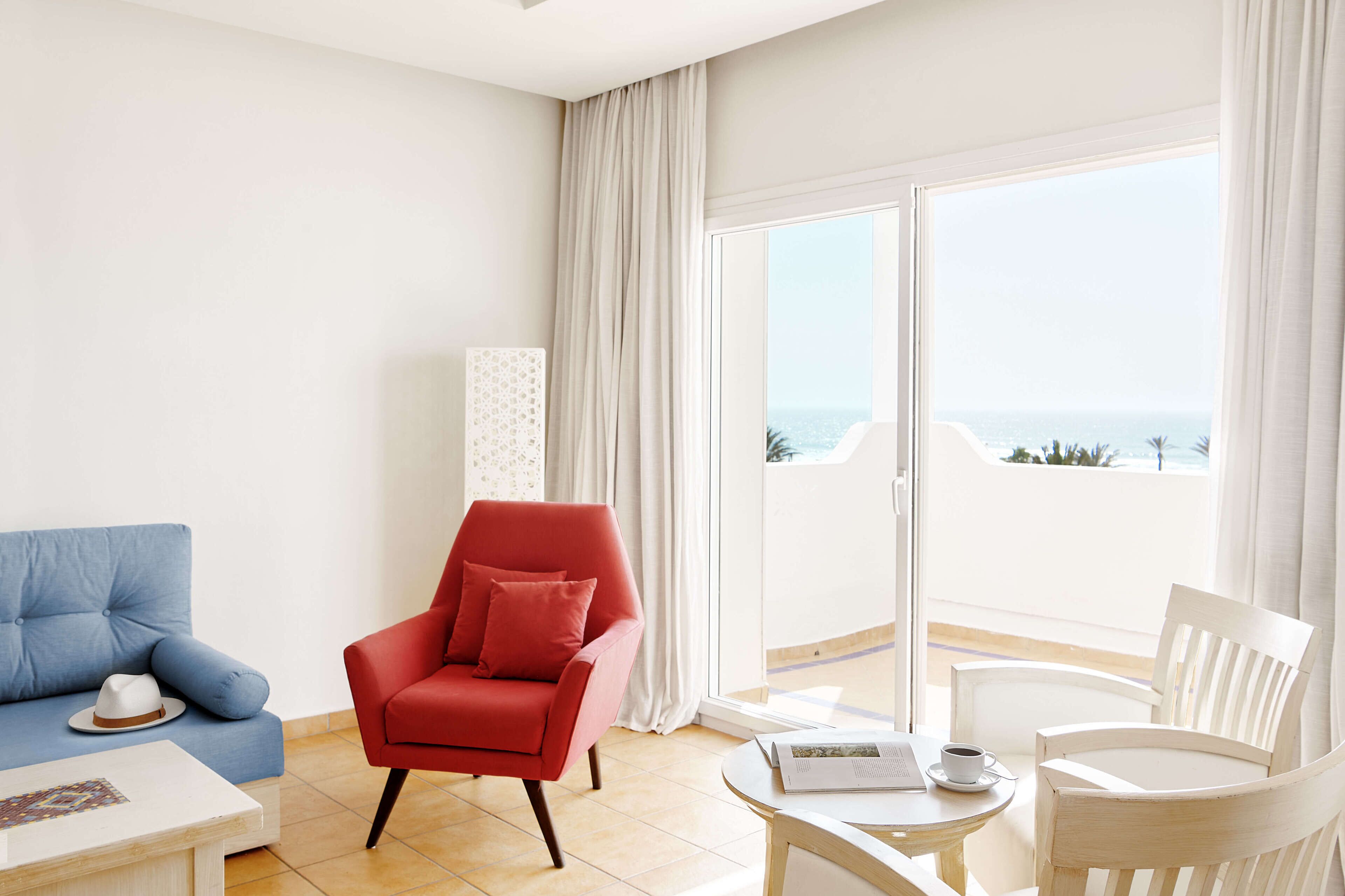 Et moderne hotelværelse med en blå sofa, en rød lænestol, en hvid lænestol og en balkon med havudsigt.