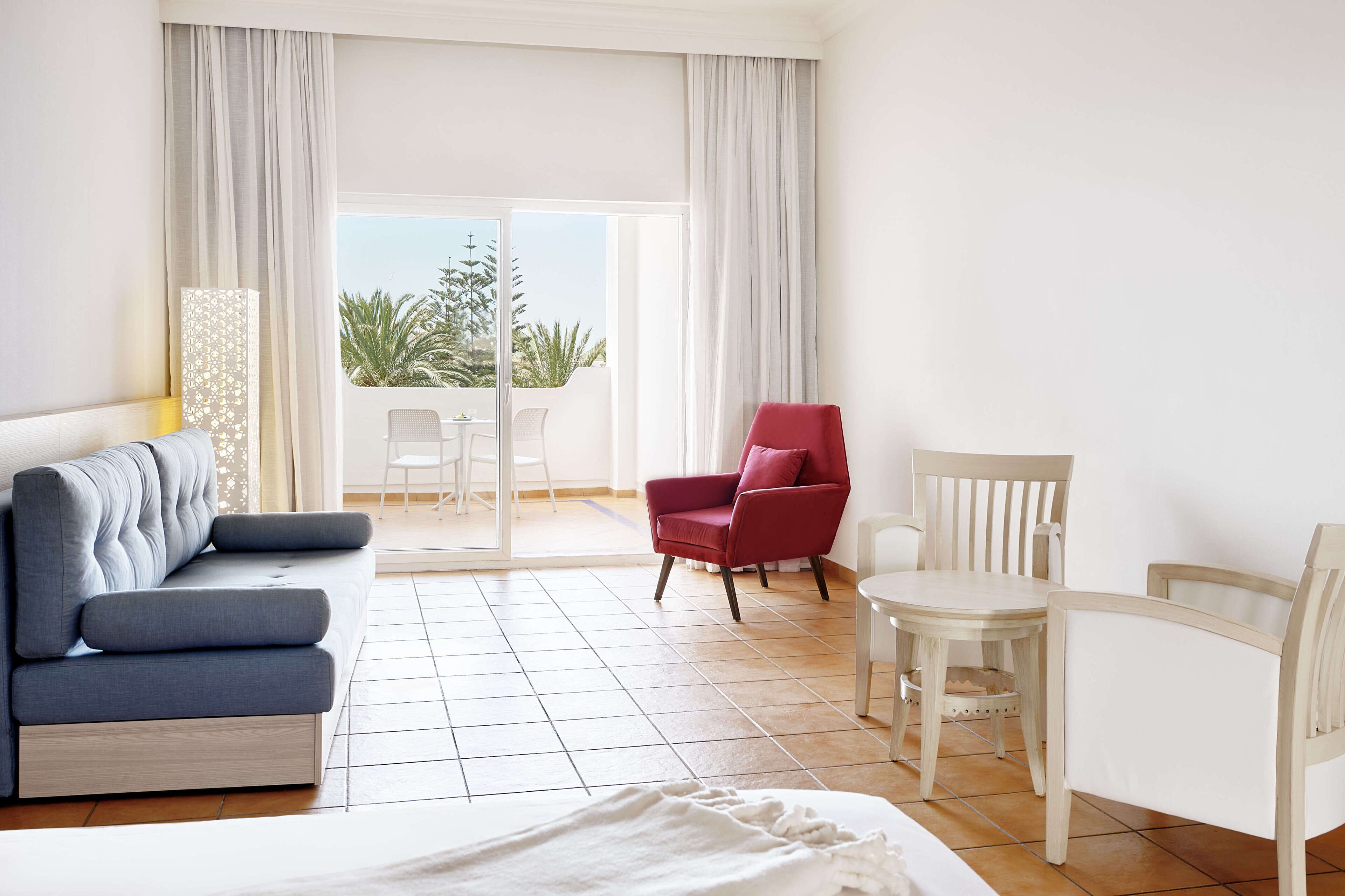 Une chambre d’hôtel avec un lit, un canapé bleu, un fauteuil rouge, une table blanche et une vue sur des palmiers, visible à travers une porte-fenêtre coulissante.