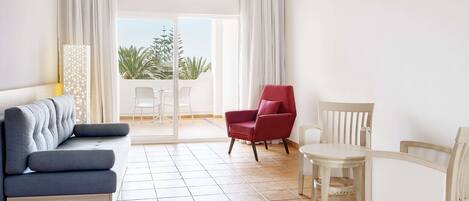 Une chambre d’hôtel avec un lit, un canapé bleu, un fauteuil rouge, une table blanche et une vue sur des palmiers, visible à travers une porte-fenêtre coulissante.