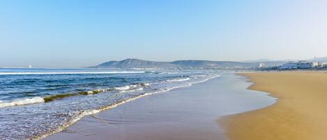 A wide sandy beach with gentle waves, a clear sky, and distant mountains.