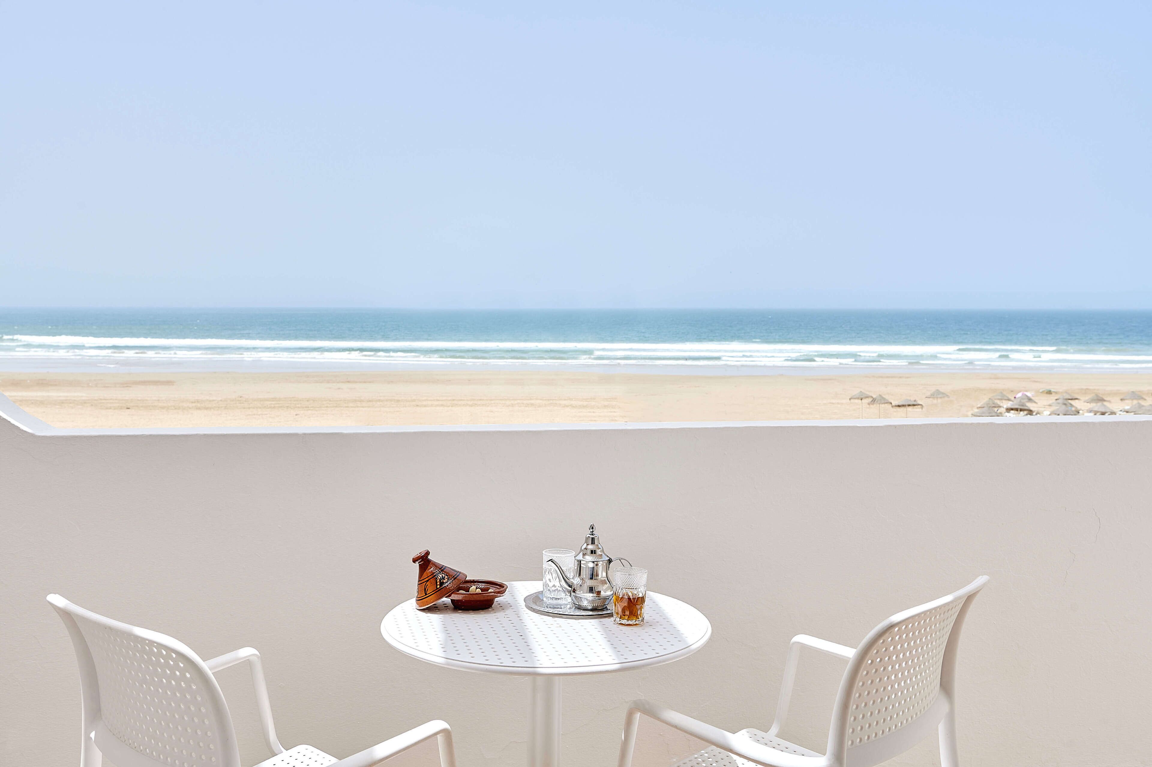 A table with a teapot, cup, and jug on a balcony overlooking a beach.