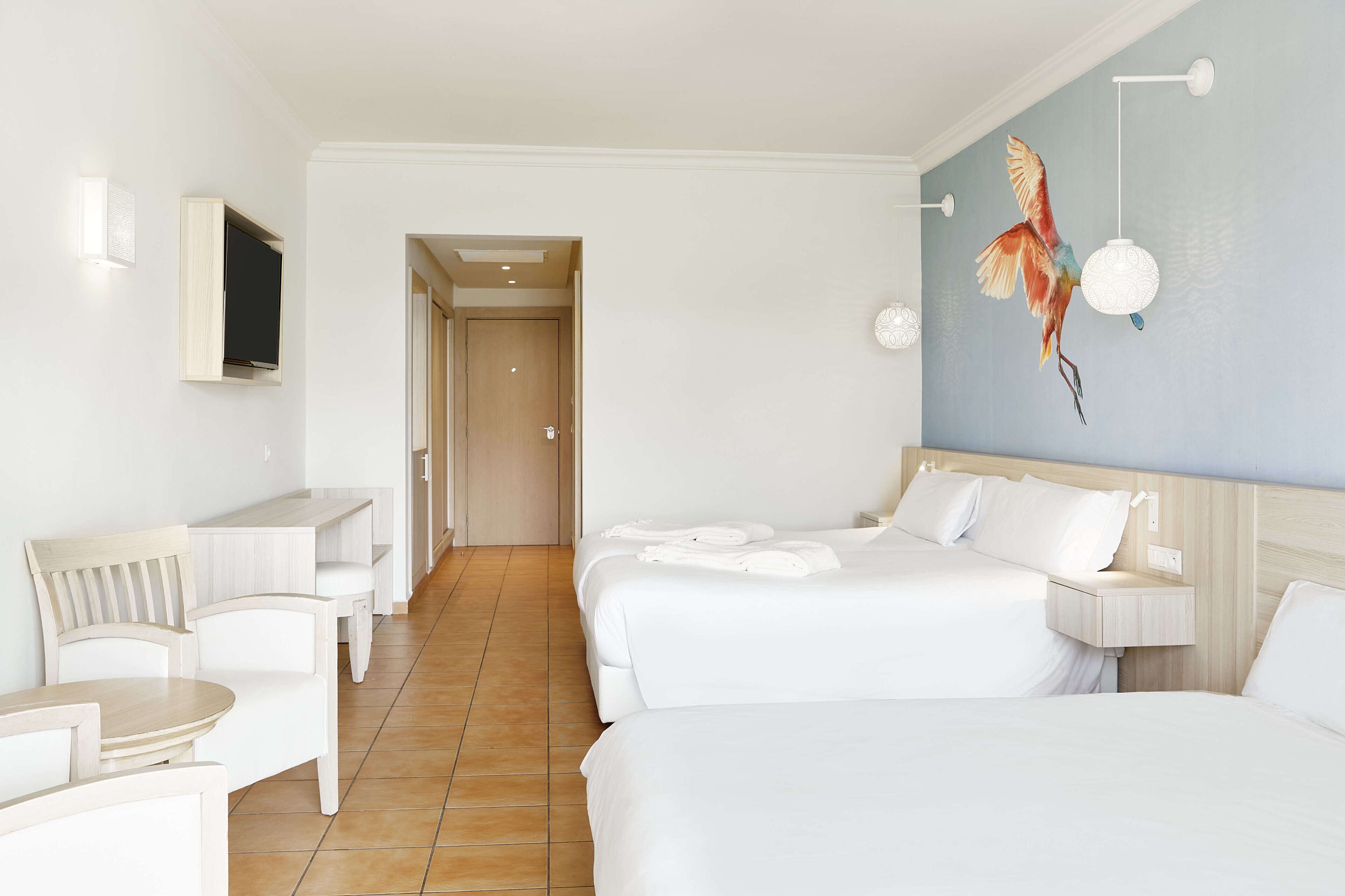 A hotel room with two beds, a TV mounted on the wall, a painting of a bird, and a small table with chairs.