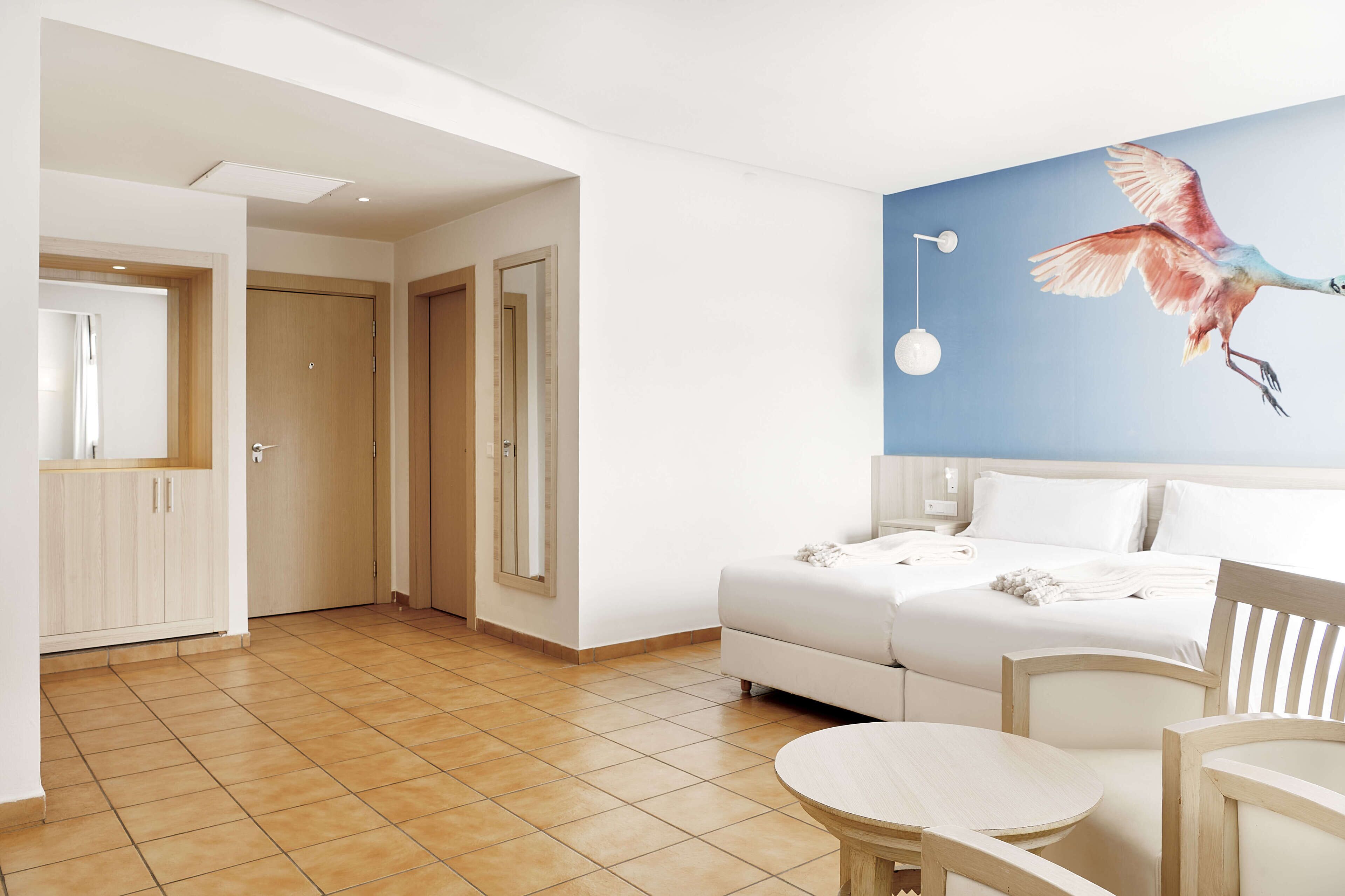 A hotel room with a bed, a dining table, chairs, a painting of a bird on the wall, and a view of the sea.